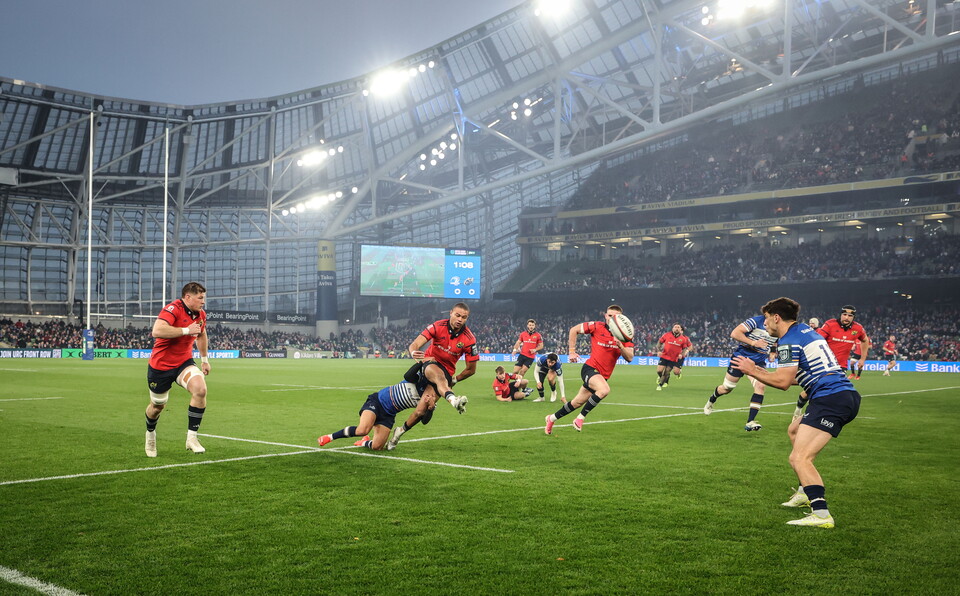 Rugby players in red and blue kits compete on a green field under stadium lights.