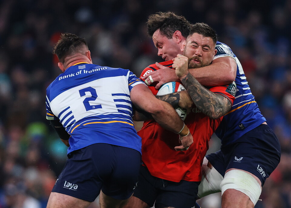 Three rugby players, one with a red jersey, one in blue and white, and another in blue and white, grappling for the ball.