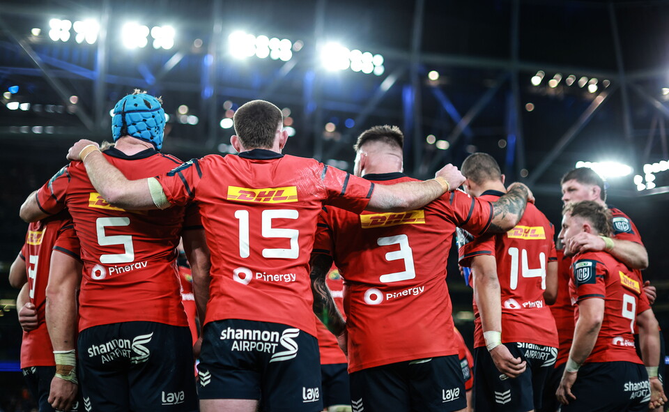 Rugby players wearing red shirts huddle on a field with numbers 5, 15, 3, 14, and 9.