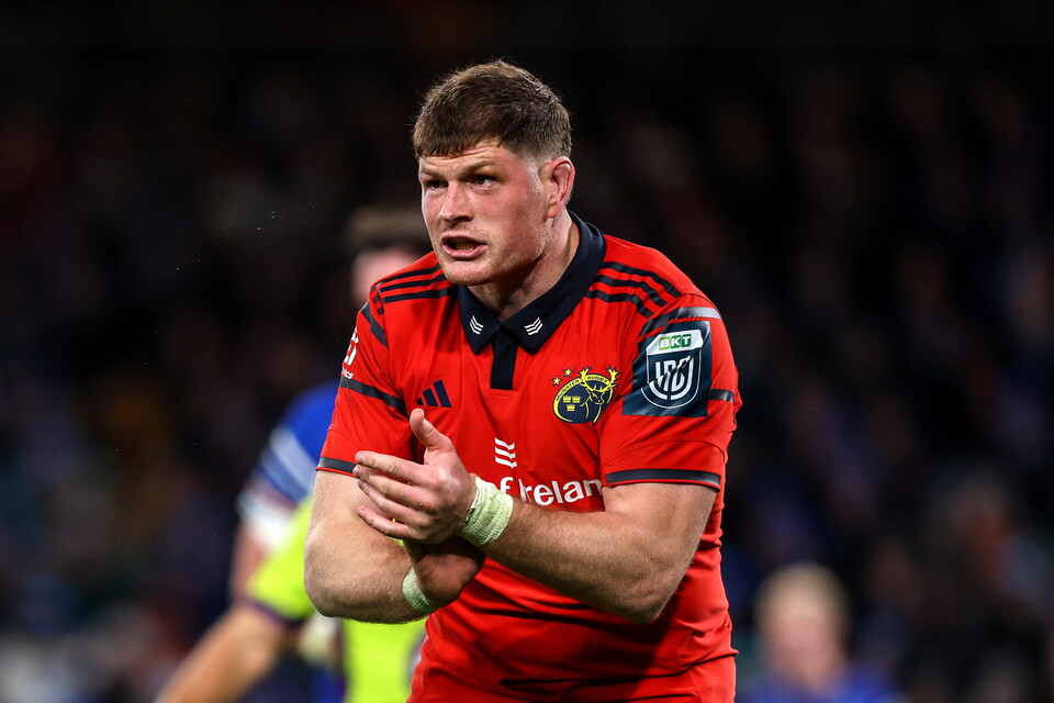 Player in red rugby shirt clapping hands, watching match, audience blurred in background.
