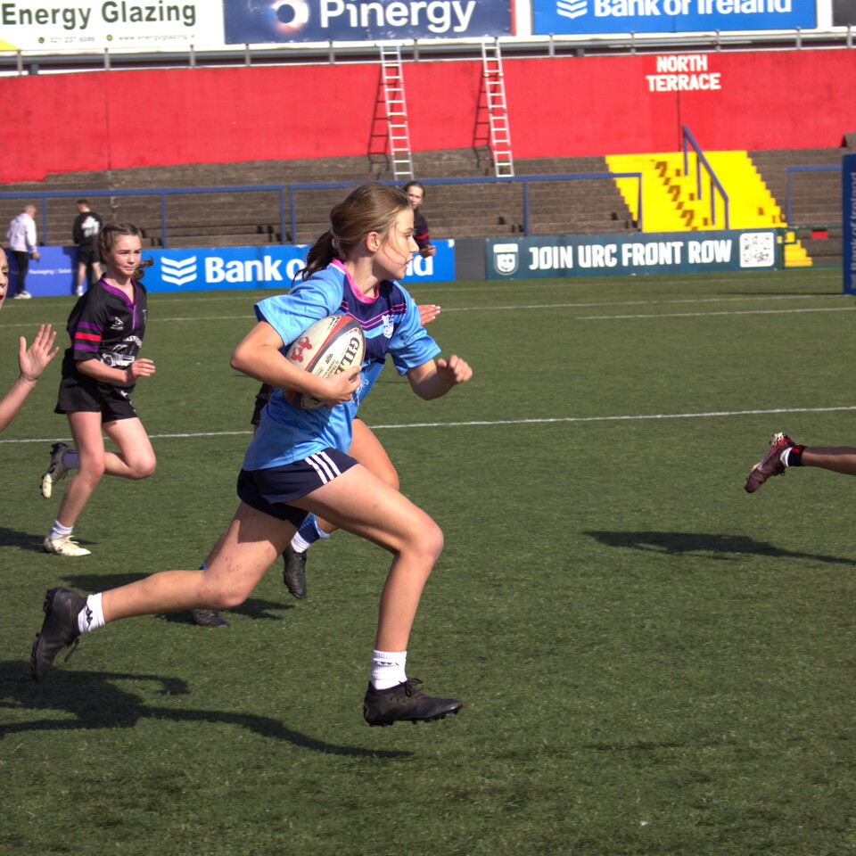 A female rugby player in a blue shirt running with the ball on a field.