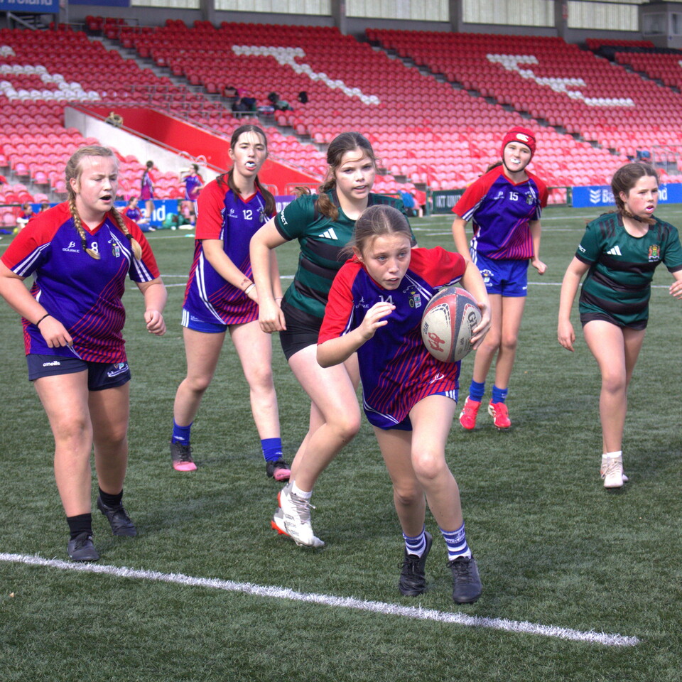 Seven girls in different uniforms play rugby on a green field.