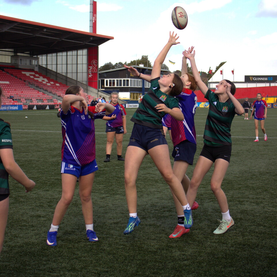 Five women players in green, blue and purple jerseys jump to catch a rugby ball.