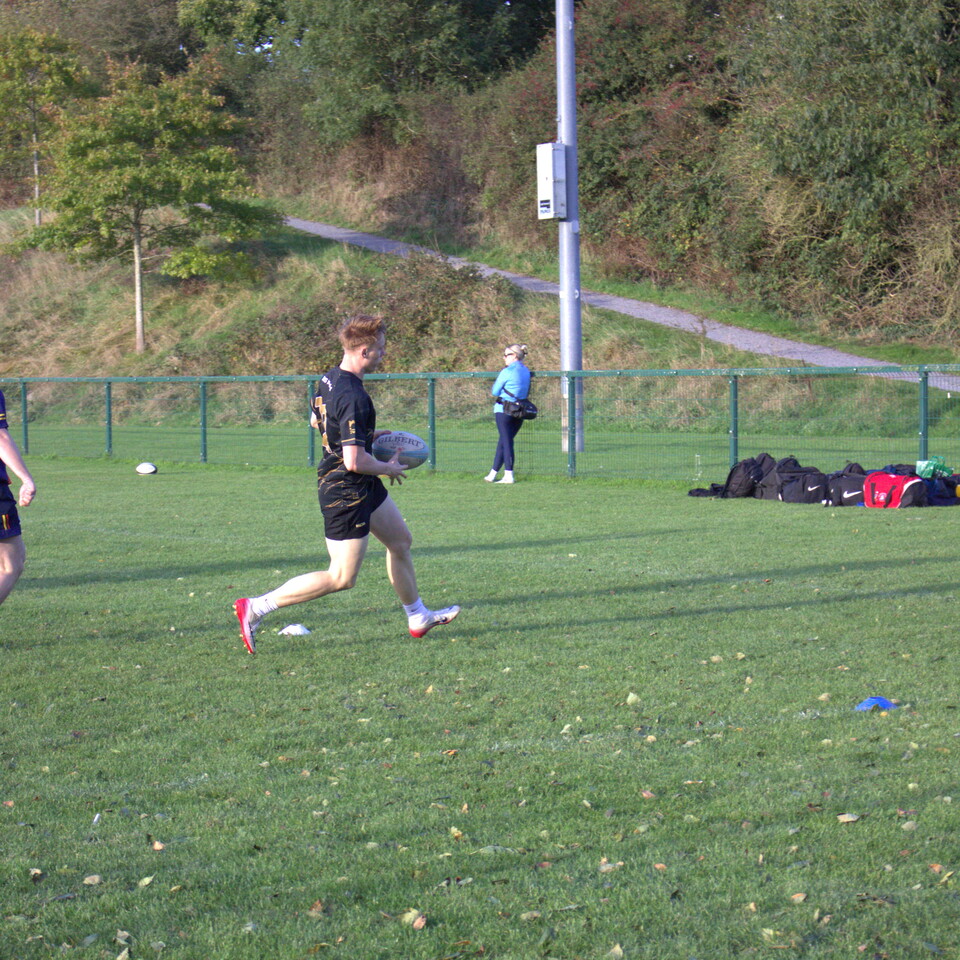 Player in black runs with rugby ball in hand, another player and bag on grass field.