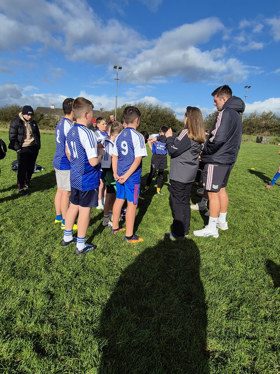 Young footballers wearing blue and white shirts gather around a coach on a grassy field.