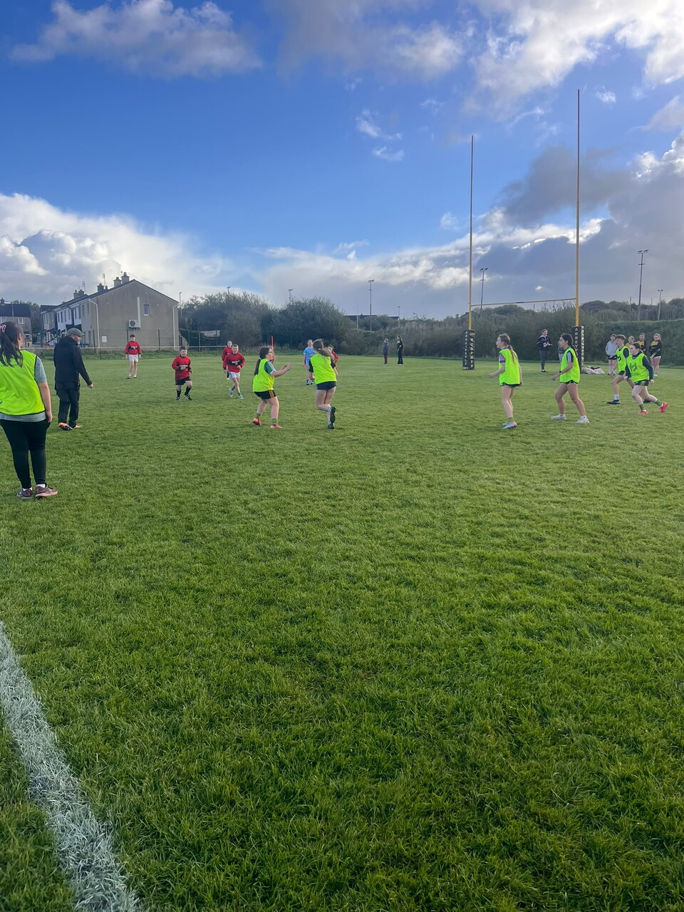 Players in yellow vests run on a green field under a partly cloudy sky.