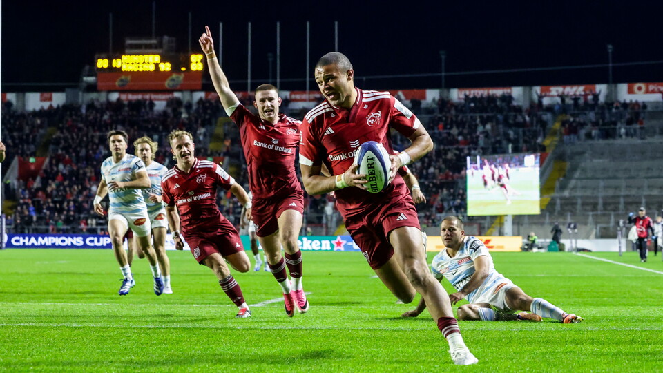 Players in red jerseys run on a rugby field with one player holding the ball.