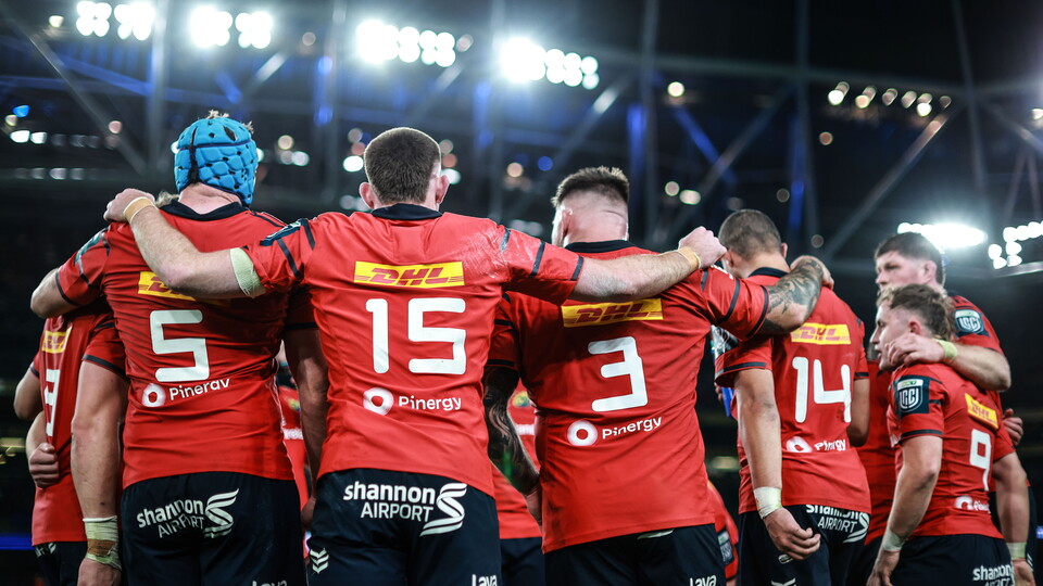 A group of players in red shirts huddle together on a rugby pitch.