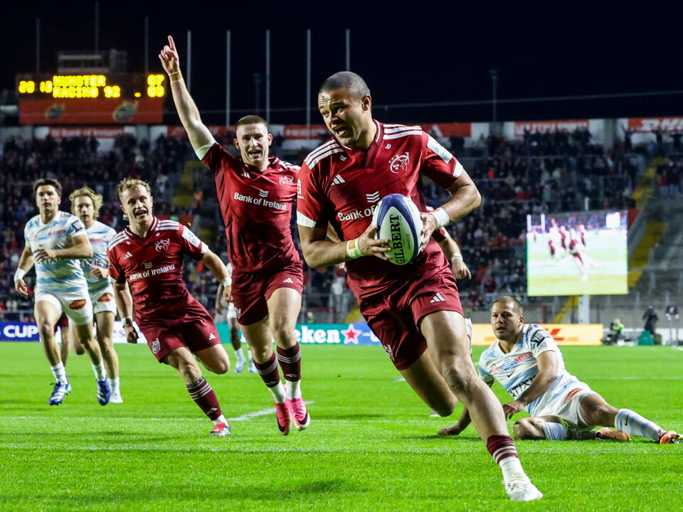 Players in red jerseys run on a rugby field with one player holding the ball.