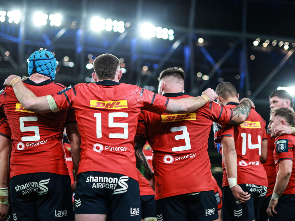 A group of players in red shirts huddle together on a rugby pitch.